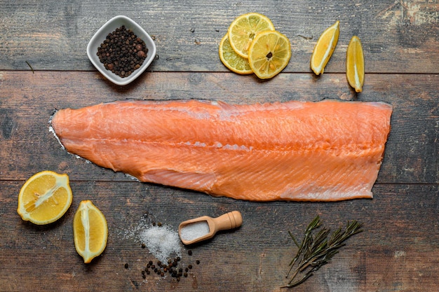 Fish fillet with culinary ingredients salt herbs pepper and lemon raw Atlantic salmon fillet on skin on dark wooden board during cooking