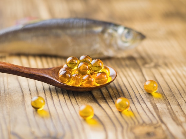 Fish oil capsules in a wooden spoon on a wooden table with a fish in the background Selective focus