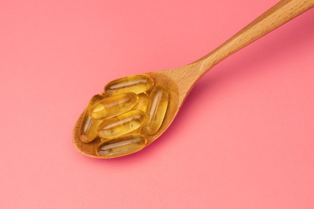 Fish oil. Wooden spoon with Omega 3 capsules on pink background. Top view.