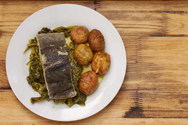 Fish with potato and vegetables on white plate and glass of water on brown