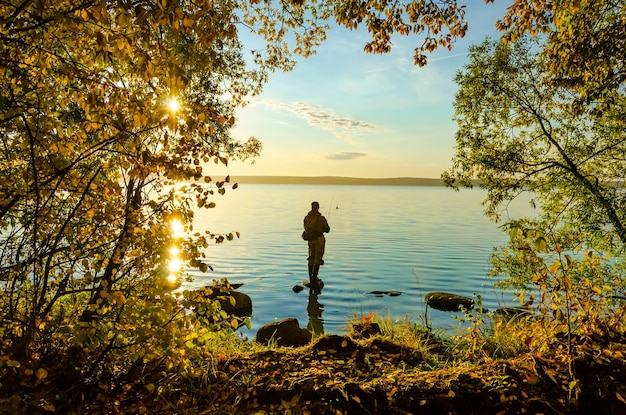 A fisherman stands on a rock in the lake near the autumn forest.