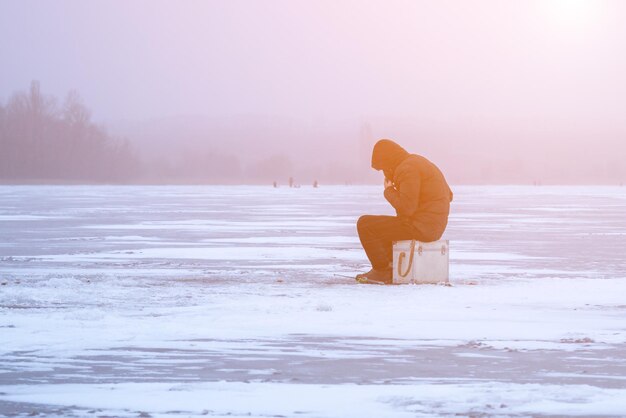 A fisherman on winter fishing on a frosty evening sits on an ice floe in the fog a winter landscape