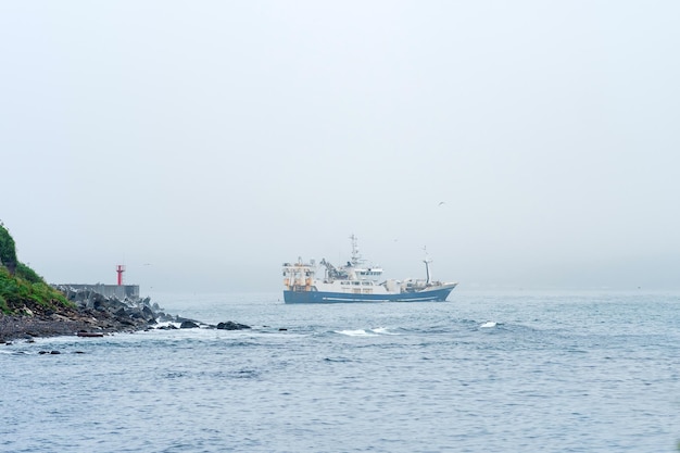 Fishing vessel emerges from behind a cape with a lighthouse sailing into a foggy sea