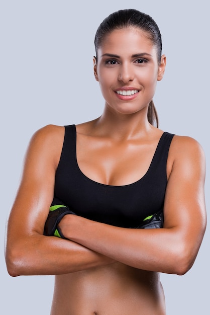 Fit and beautiful. Beautiful young sporty woman in sports clothing keeping arms crossed and smiling while standing against grey background