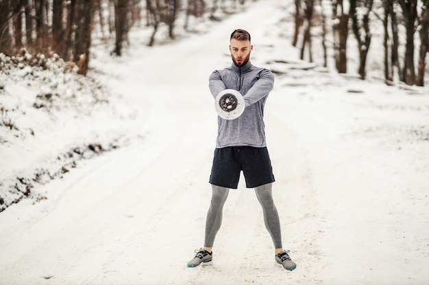 Fit bodybuilder standing on snowy path in forest and swinging kettlebell at winter. Bodybuilding exercises, winter fitness, outdoor fitness