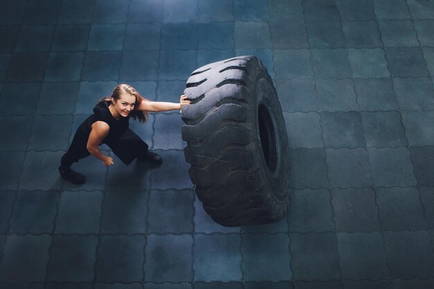 Photo fit female athlete working out with a huge tire turning and carry in the gym woman exercising with