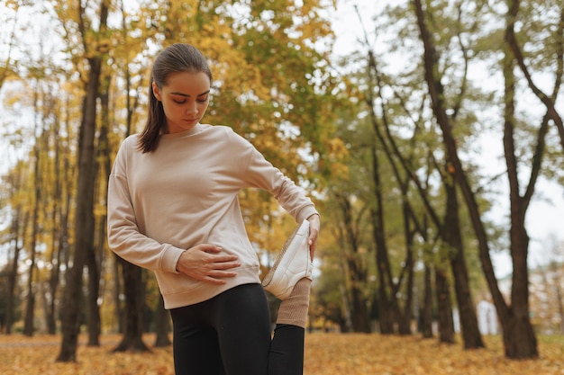 Fit female runner stretching leg while standing in autumn park and warming up during training