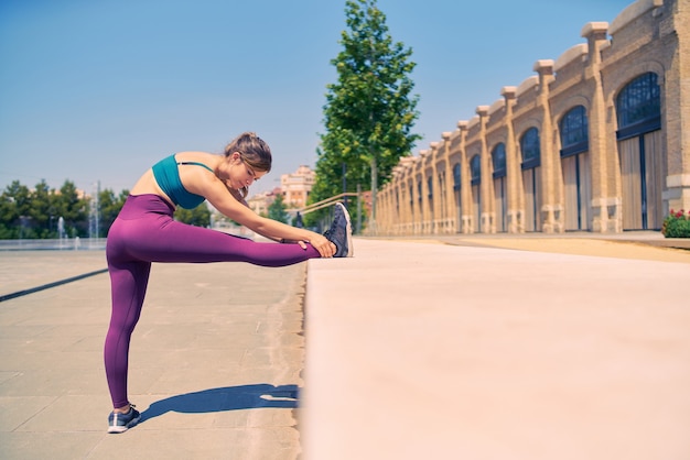 Fit girl stretching legs in a city park wearing vibrant purple sportswear and blue bra healthy lifestyle