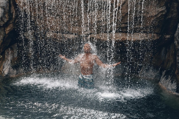 Fit and handsome topless Arabic man under the waterfall, huge rocks and water, splashes and flecks; natural beauty concept.