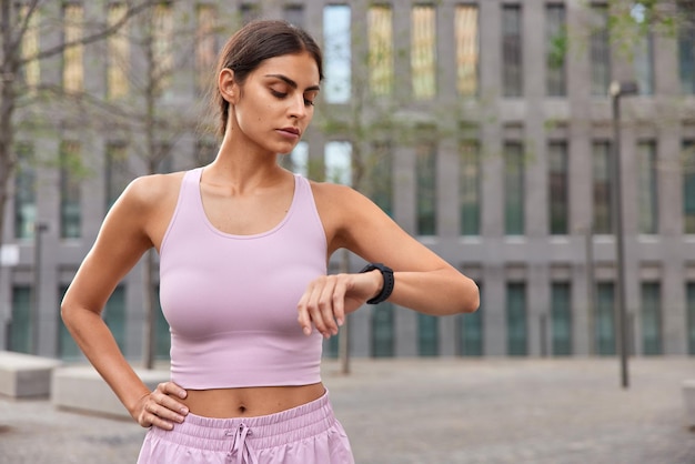 Fit healthy brunette young woman dressed in sportswear checks fitness results via smartwatch keeps hand on waist goes in for sport outdoors during summer time poses against modern city building