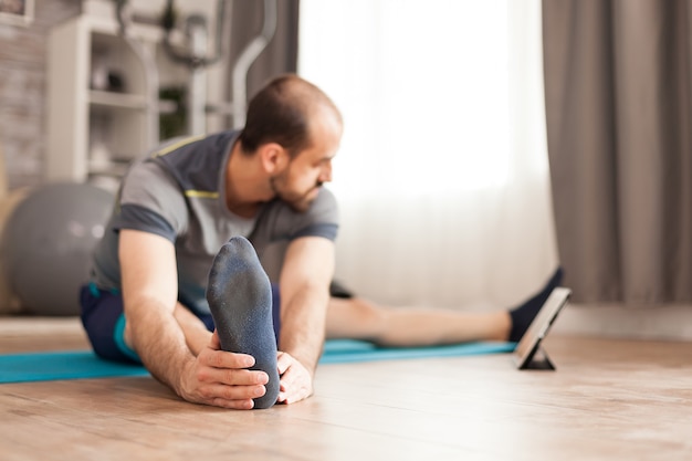 Fit male stretching his body watching online pilates class on tablet computer during global lockdown.