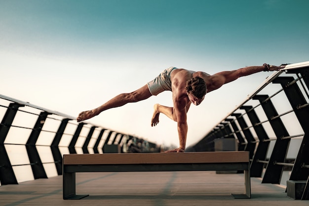 Fit man doing a one hand stand while practicing yoga alone near the ocean against sky at dusk or dawn