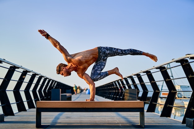 Fit man doing a one hand stand while practicing yoga alone near the ocean against sky at dusk or dawn