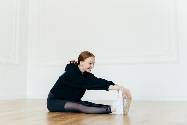 Fit redhead woman stretches muscles on legs has sporty body focused into distance looks happily wears black clothes poses on floor in spacious hall People flexibility and sport concept