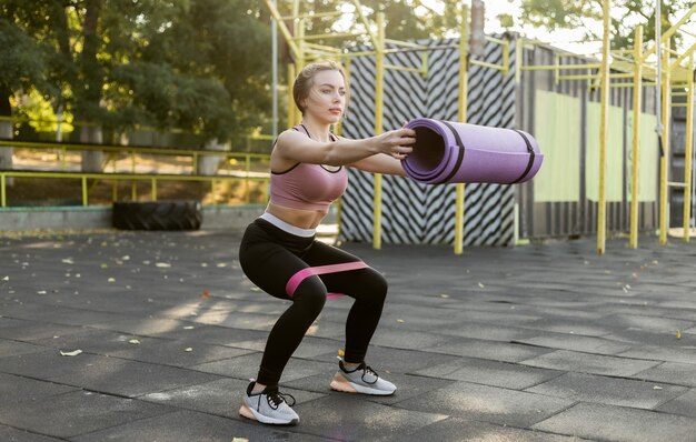 Fit slim woman exercises legs with fitness rubber bands and holding mat at the sports ground