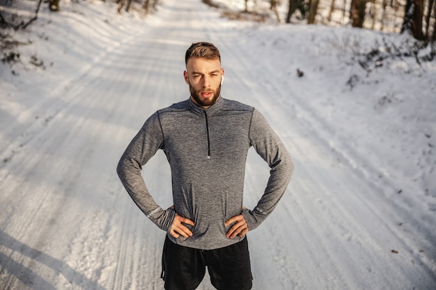 Fit sportsman standing on snowy path in nature and holding hands on hips. Winter sport, winner, achievement