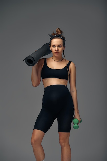 Fit young woman exercising during her pregnancy standing smiling at the camera with bottled water in one hand and a gym mat in the other