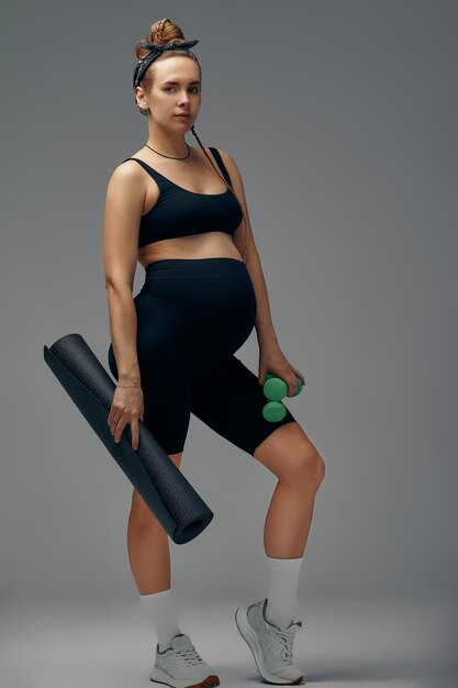 Fit young woman exercising during her pregnancy standing smiling at the camera with dumbbells in one hand and a gym mat in the other