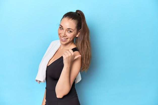 Fit young woman with a towel on her shoulder studio shot points with thumb finger away