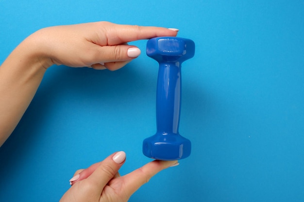 Fitness concept A female hand holds a blue plastic dumbbell Blue background