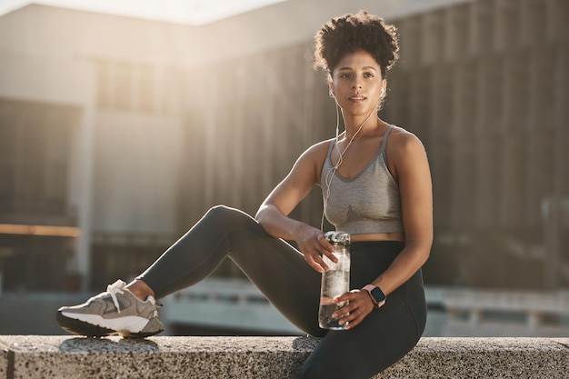 Fitness earphones and black woman water bottle break and training workout exercise and motivation in urban city Thinking young athlete sports hydration and nutrition for body wellness and goals