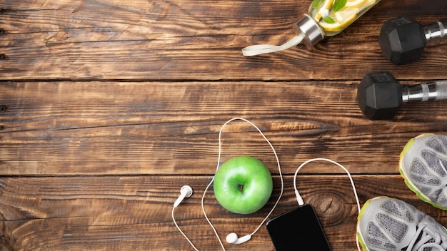 Fitness equipment on wooden background with copy space: dumbbells, phone with headphones, green apple, sneakers.