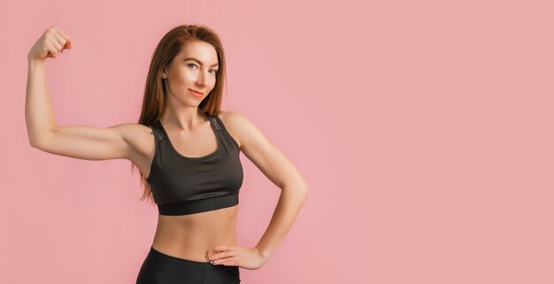 Fitness girl smiling in black sportswear on a pink background. Slim woman with a beautiful athletic body and tanned skin