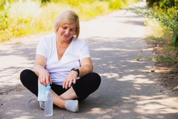 Fitness happy woman sitting in the lotus position on the road in the park single with a bottle of water down, keeping medical mask on her hand. High quality photo