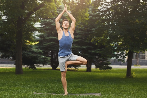 Fitness, man training yoga in tree pose outdoors. Young sporty guy makes breathing exercise in park, copy space