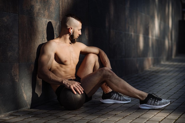 Fitness muscular man with weight ball resting and sitting against the concrete wall