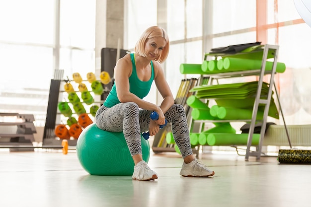 Fitness smiling woman resting with dumbbells while sitting on fitness ball in light gym Healthy lifestyle