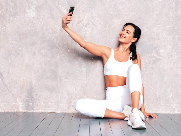 Fitness smiling woman in white sports clothing Sexy young beautiful model with perfect body Female posing near grey wall in studio Stretching out before training Taking selfie photos