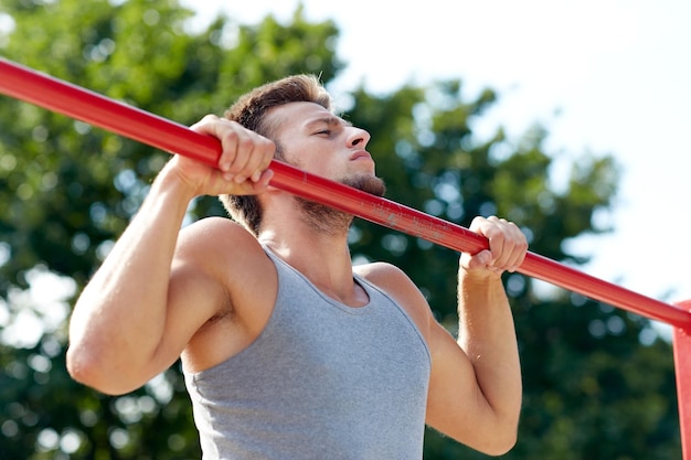 fitness, sport, exercising, training and lifestyle concept - young man doing pull ups on horizontal bar outdoors