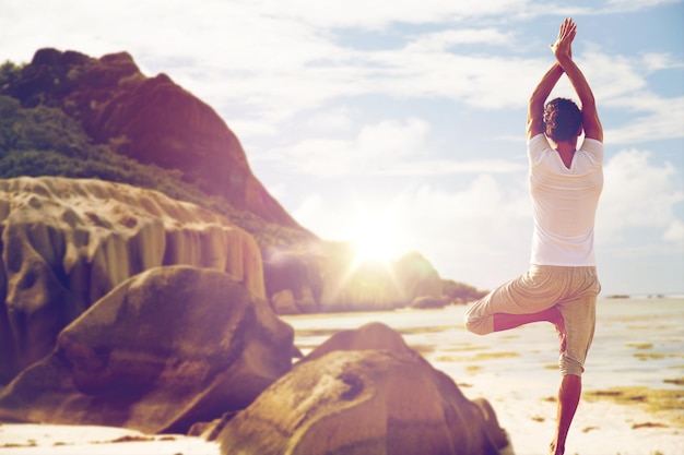 fitness, sport, people and healthy lifestyle concept - man meditating in yoga tree pose over exotic beach background from back