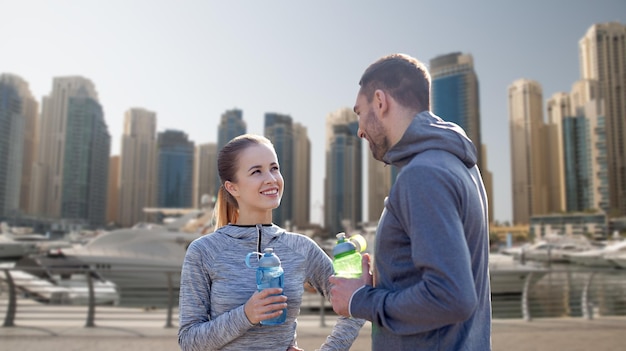 fitness, sport, people and lifestyle concept - smiling couple with bottles of water outdoors over dubai city street background