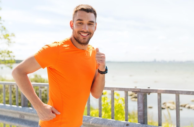 fitness, sport, people, technology and healthy lifestyle concept - smiling young man with heart rate watch running at summer seaside