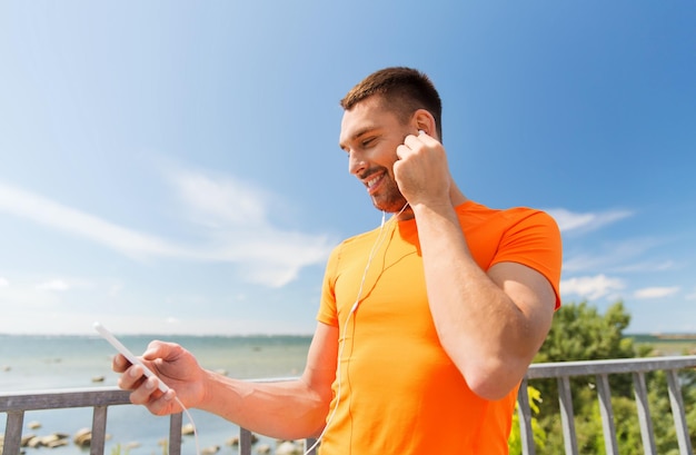 fitness, sport, people, technology and healthy lifestyle concept - smiling young man with smartphone and earphones listening to music at summer seaside