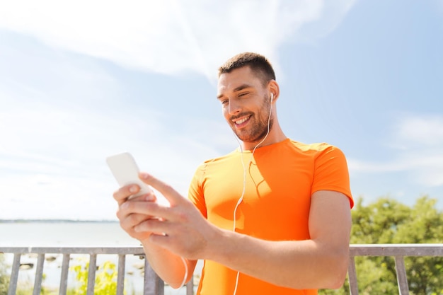 fitness, sport, people, technology and healthy lifestyle concept - smiling young man with smartphone and earphones listening to music at summer seaside