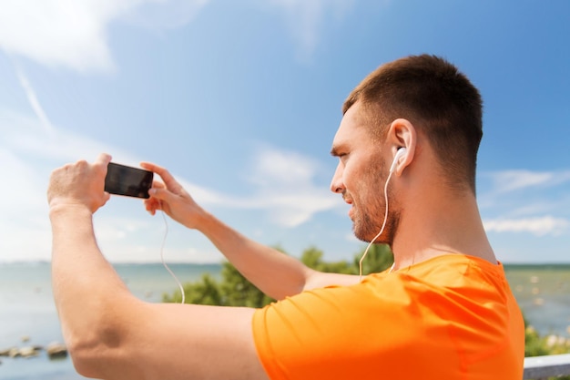 fitness, sport, people, technology and healthy lifestyle concept - smiling young man with smartphone and earphones listening to music at summer seaside