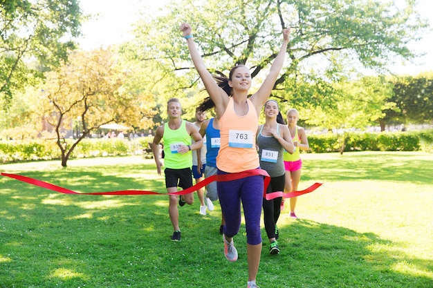 fitness, sport, victory, success and healthy lifestyle concept - happy woman winning race and coming first to finish red ribbon over group of sportsmen running marathon with badge numbers outdoors