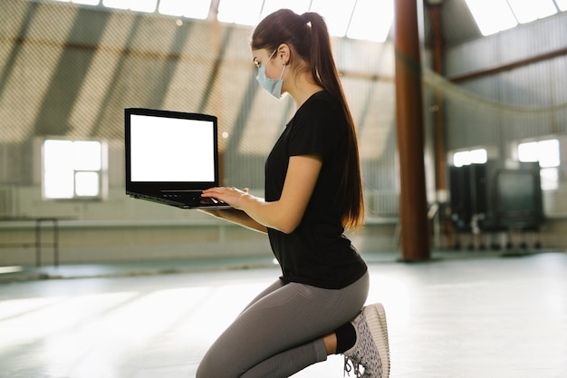 Fitness trainer kneel down in empty gym woman in medical mask works remotely indoor in solitude she sits with laptop on knees lockdown quarantine of coronavirus covid self isolation