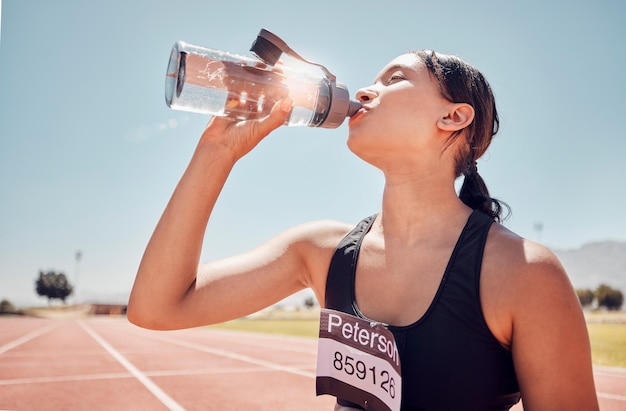 Fitness water and runner woman training at running track thirsty and relax after workout against a blue sky background Sports girl and drinking water at stadium after running exercise and sport