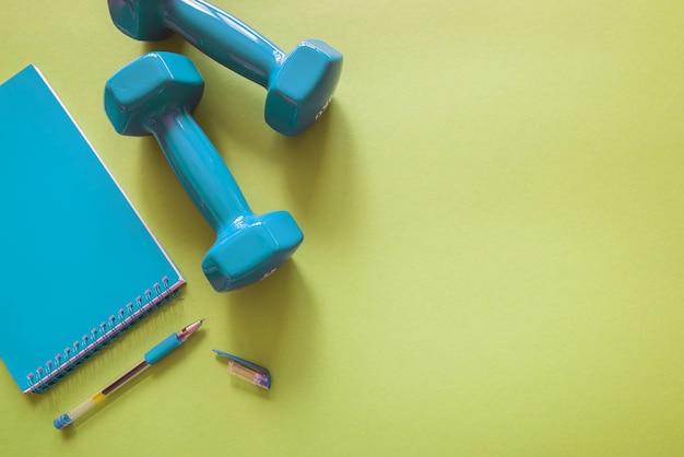 Fitness and weight loss concept Blue dumbbells with notepad on green background with copy space