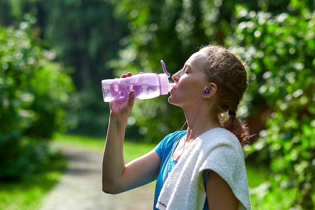 Fitness woman drinking water after running training in summer park.