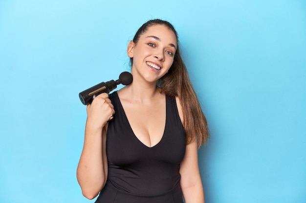 Fitness woman holding electric massager in a blue studio setting