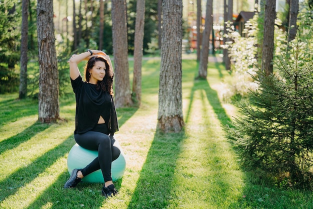 Fitness woman looks into distance with thoughtful expression sits on fitness ball wears black t shirt and leggings poses at green spring forest keeps arms raised over head has workout outdoor