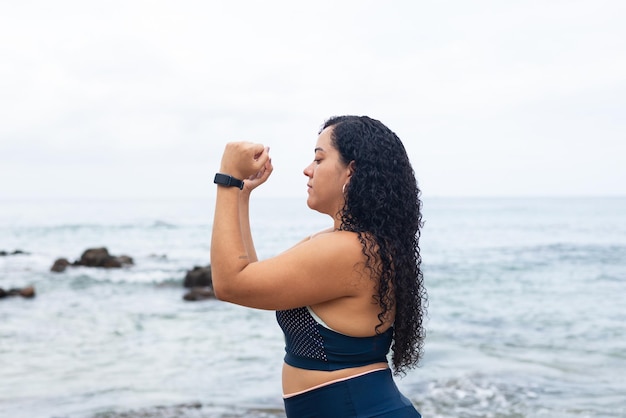 Fitness woman standing doing exercises with her arms on the beach sand against the sea and cloudy sky