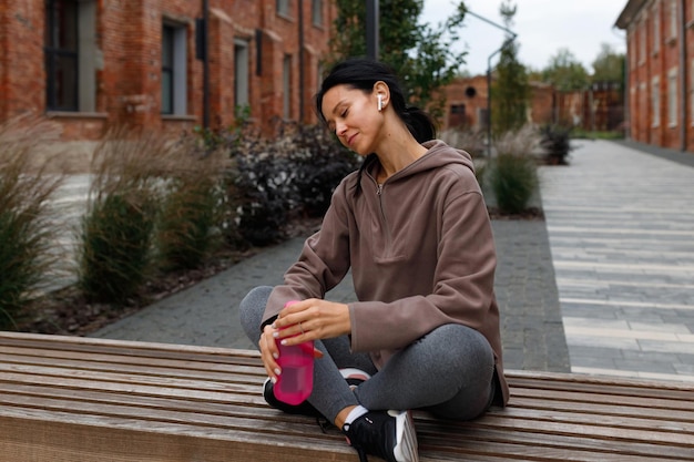 Fitness woman in wireless headphones and sports bottle in hands sits on bench