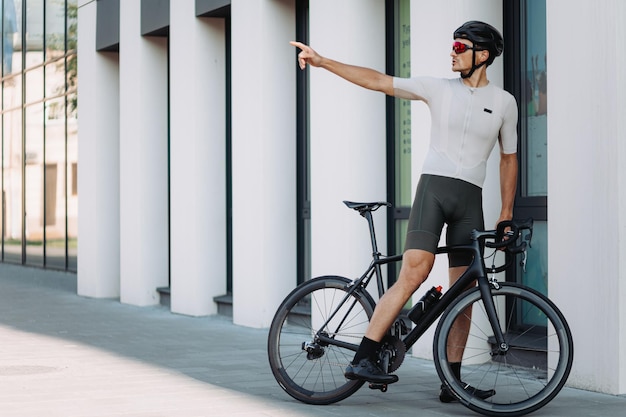 Fitness young man in safety helmet and glasses sitting on black bike and pointing with hand aside Outdoors workout of caucasian cyclist Urban area