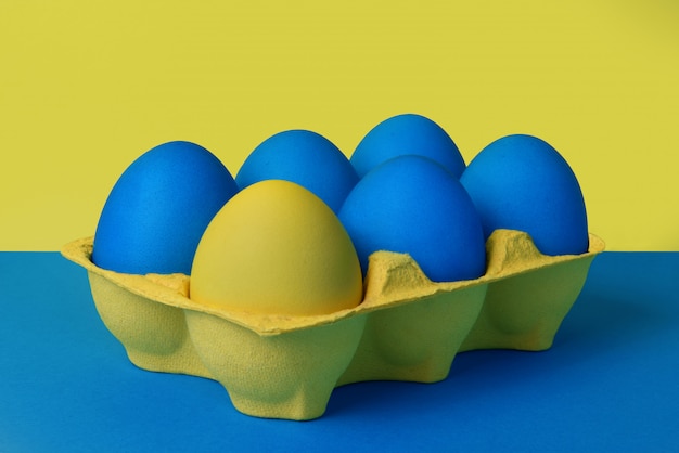 Five blue and one yellow painted Easter eggs in yellow wrapping on blue and yellow background